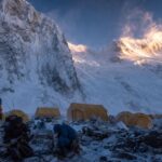 Mountaineers prepare beside tents at an advanced base camp beneath snow-covered Himalayan peaks.