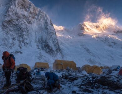 Mountaineers prepare beside tents at an advanced base camp beneath snow-covered Himalayan peaks.