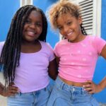Two smiling young girls standing beside a bright blue building in casual summer clothes.
