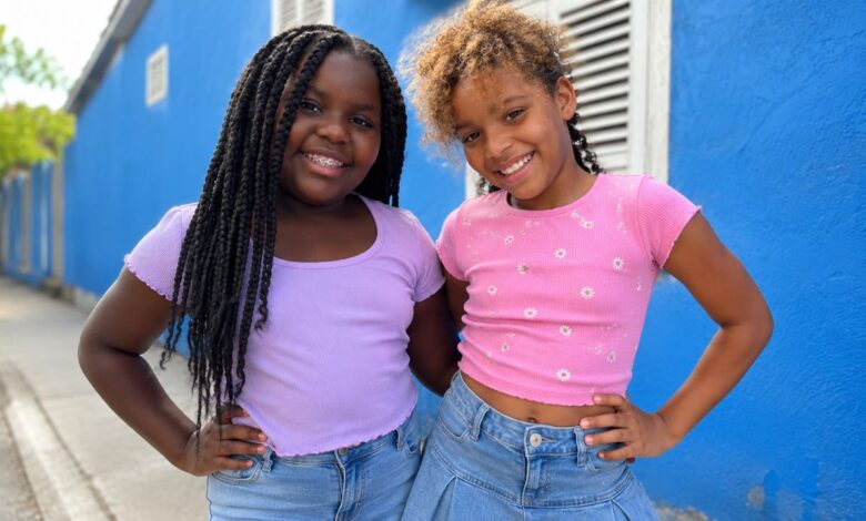 Two smiling young girls standing beside a bright blue building in casual summer clothes.