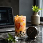 An elegant transparent glass of modern fruit-and-milk tea with tapioca pearls, placed next to a minimalist analog clock and a blurred laptop